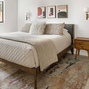 A modern bedroom with a neatly made bed featuring beige bedding and pillows. The room has white walls with three abstract art pieces above the bed, including a touch of Eichler Orange, and a wooden nightstand with a wall-mounted sconce.