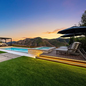 Backyard view with a pool, hot tub, and seating area under a pergola, set against the majestic Sunset Trails mountainous landscape at dusk.