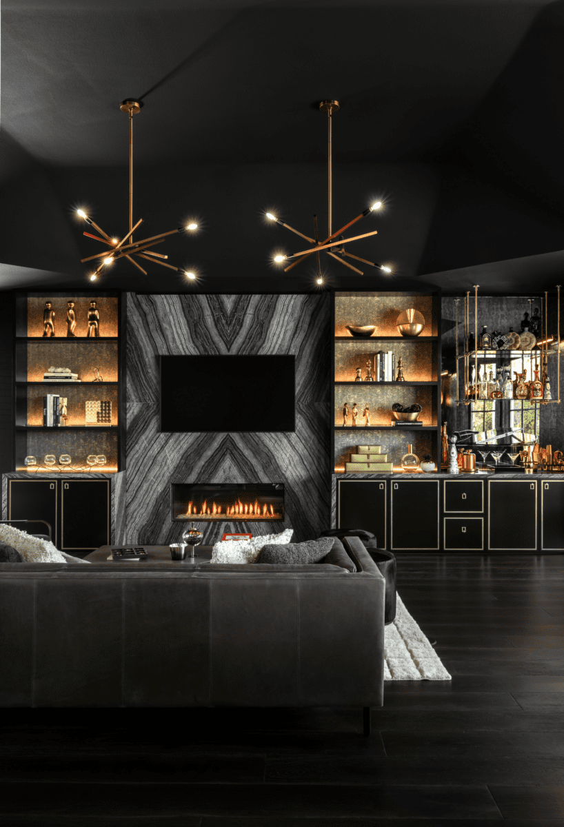 Modern living room with sophisticated glamour: a large sofa faces a wall-mounted TV above a fireplace, while open shelves showcase various decorations and a built-in bar area. The ceiling features two elegant pendant lights.