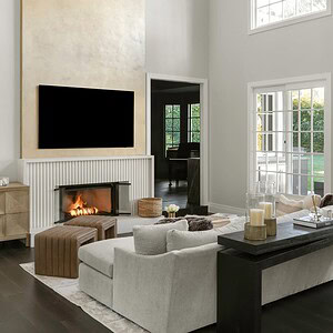 Modern living room with high ceilings, neutral color palette, a large TV above a fireplace, and a sectional sofa. Large windows allow natural light in, enhancing the room's sophisticated glamour.