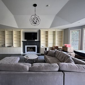 A living room exuding sophisticated glamour, featuring a large gray sectional sofa, a coffee table, and a fireplace with a TV above it. Built-in shelves add charm, while boxes and other items are seen near the windows in the background.
