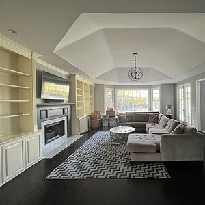 A spacious living room with a sectional sofa, coffee table, built-in shelves, a fireplace, and a large TV exudes sophisticated glamour. The room features a zigzag patterned rug, contemporary artwork, and large windows that flood the space with natural light.
