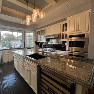 A modern kitchen exuding sophisticated glamour with white cabinets, stainless steel appliances, a large island, and pendant lights. The space includes a sink, dishwasher, oven, and wine cooler, complemented by large windows that flood the room with natural light.