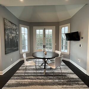 A modern dining area with a touch of sophisticated glamour features a round table and four chairs, a large painting on the wall, French doors leading outside, a wall-mounted TV, and an area rug covering dark hardwood floors.