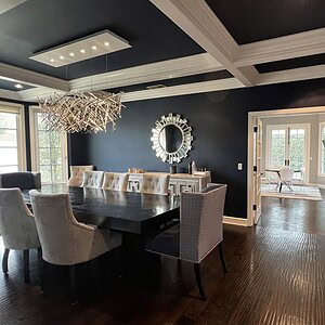 A dining room with sophisticated glamour, featuring a dark wooden table, upholstered chairs, a large mirror on the wall, and an abstract painting. A modern chandelier hangs above the table, while an adjoining room can be seen in the background.