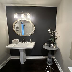 Modern bathroom with a black accent wall exudes sophisticated glamour. Includes a pedestal sink, round mirror with decorative frame, wall lighting, small table with plants, and a framed photo of a baby.