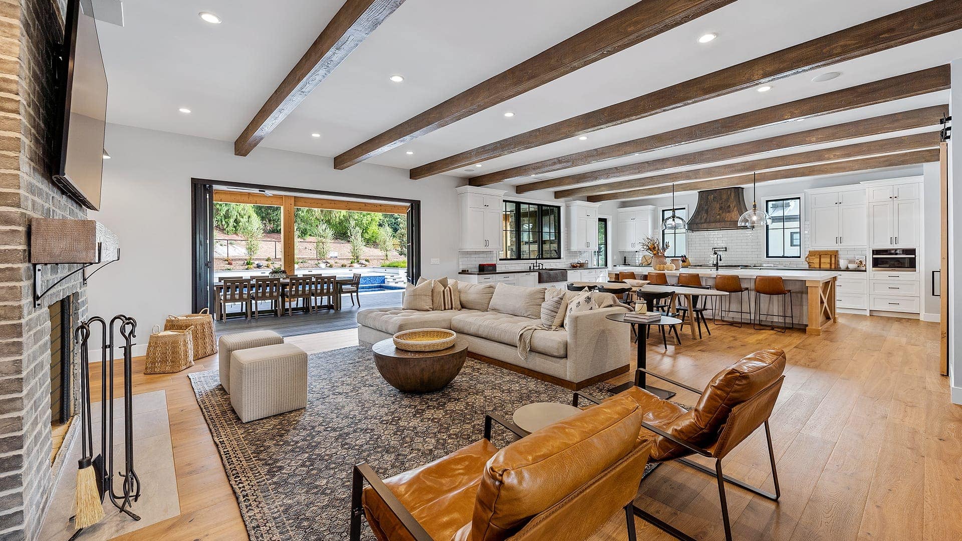 Modern open-concept living room and kitchen with wooden ceiling beams, light wood flooring, leather chairs, and a large rug. Glass doors open to an outdoor dining area with a pool—what makes us different is the seamless blend of indoor comfort and outdoor luxury.