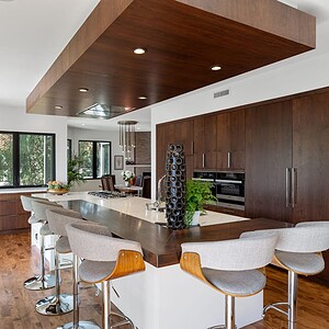 Modern kitchen with wooden cabinets, a central island with bar stools, white countertops, recessed lighting, and large windows providing natural light. Green plants are placed around the space for a touch of modern refreshment.