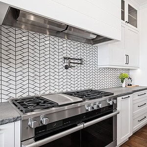 Modern kitchen featuring a Carrita Seaview stainless steel stove and oven, white cabinets with black handles, a chevron-patterned tile backsplash, and wooden flooring.
