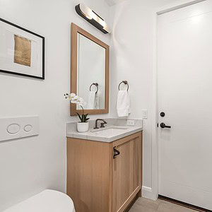 A modern bathroom with light wood cabinets, white walls, and a towel ring holding a white towel. Adorned with artwork on the wall and a white door, it features a sink with a faucet, an orchid plant, and an overhead light fixture. The Carrita Seaview theme adds an elegant touch to the serene space.