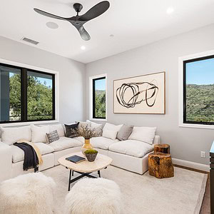 A modern living room with large windows overlooking Carrita Seaview. It features a white sectional sofa, a minimalist coffee table, two white ottomans, and wall art. There is a ceiling fan and a TV on the right.