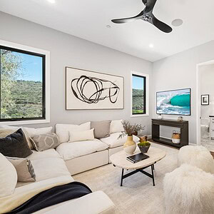 A modern living room with a white sectional sofa, a TV displaying a Carrita Seaview beach scene, abstract wall art, a wooden coffee table, and two fluffy stools. The room has two large windows and a ceiling fan.