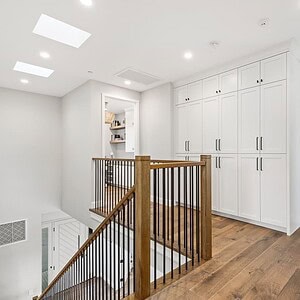 A bright hallway featuring wooden flooring and a staircase with a dark wooden handrail. The walls are painted white, and there are white built-in storage cabinets. A room with windows, offering a glimpse of the Carrita Seaview, is visible in the background.