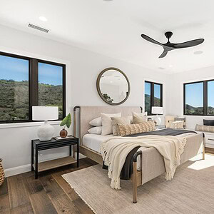 A modern bedroom at Carrita Seaview features a beige bed with pillows and blankets, a large round mirror, three windows with scenic views, a ceiling fan, and contemporary furniture on a wooden floor.