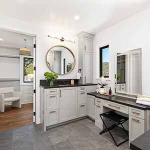 Modern bathroom with gray cabinetry, round mirror, black countertops, and two windows featuring a stunning Carrita Seaview. A vanity area and adjoining room with a white chair are visible.
