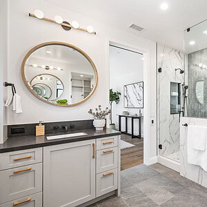 Modern bathroom with a glass-enclosed shower, marble tiles, a round mirror above a vanity with gray cabinets, and a view into an adjacent room with framed Carrita Seaview wall art.