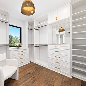 Spacious walk-in closet with open white shelving, a white upholstered chair, a window offering views of Carrita Seaview, wooden floor, a pendant light, and floral decor.