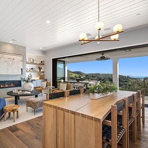 A modern living room with wood flooring, a fireplace, and artwork on the wall. The dining area connects seamlessly to large glass doors opening onto an outdoor patio with scenic hilltop views of Carrita Seaview.