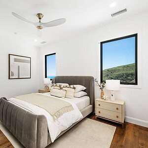A modern bedroom features a grey upholstered bed, nightstands with lamps, and a large painting on the wall. A door opens to a bright bathroom. The window offers a view of the green hillside and beyond it, the breathtaking Carrita Seaview.