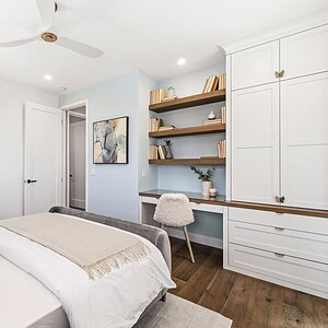 A modern bedroom with white walls and built-in shelving offers a serene retreat. The room features a bed with beige bedding, a ceiling fan, and a desk with a chair in front of shelves filled with books and decorative items, all reminiscent of the tranquility found at Carrita Seaview.