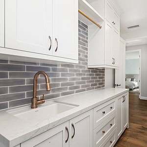 A modern laundry room at Carrita Seaview features white cabinetry, a marble countertop, a copper faucet, a tiled backsplash, and a washer and dryer beside the counter on a wooden floor.