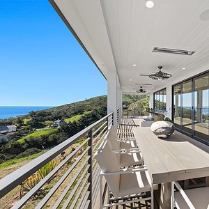 A modern outdoor deck at Carrita Seaview features a white dining table and chairs overlooking a scenic hillside and ocean. The deck includes ceiling fans and sliding glass doors leading to the interior.