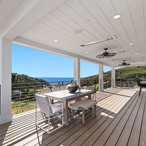 A spacious outdoor deck with a dining table and chairs overlooks the lush Carrita Seaview landscape and the ocean in the distance, under a white wooden roof with ceiling fans.