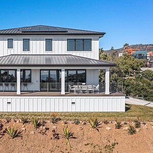 A modern two-story white house with large windows, a spacious balcony, and solar panels on the roof, situated on a hillside with surrounding greenery and offering stunning Carrita Seaviews.