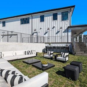Outdoor seating area with white and black furniture on a grassy lawn, adjacent to the modern Carrita Seaview two-story white building with stairs and a patio. Clear blue sky in the background.