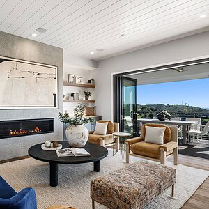 A modern living room featuring a navy sofa, beige armchairs, and a central black round table, complemented by built-in shelves with decor. A wall-mounted fireplace adds warmth, while large sliding doors open to a balcony that boasts the stunning Carrita Seaview.