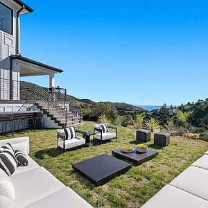 Outdoor patio area with white cushioned furniture, black coffee tables, and striped pillows on a grassy lawn overlooking the rolling green hills and distant Carrita Seaview under a clear blue sky.