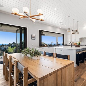 A modern kitchen and dining area with wooden floors, a large dining table, white cabinets, a gray island, and a view of the Carrita Seaview through large windows and a sliding glass door.