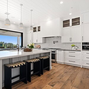 Modern kitchen with white cabinets, large marble island, three black stools, hardwood floors, stainless steel appliances, and an open window view of the scenic Carrita Seaview landscape.