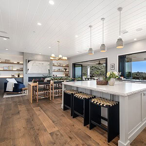 The Carrita Seaview modern, open-concept kitchen boasts white cabinetry, a large island with seating, pendant lighting, wooden flooring, and floor-to-ceiling windows offering a stunning view of the outdoor patio and ocean.