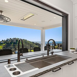 A modern kitchen sink with a large window opens to a spacious patio with outdoor seating, offering a scenic view of the hilly Carrita Seaview landscape and the ocean in the background.