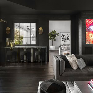 Sophisticated glamour defines this modern living room with dark walls and ceiling, featuring a chic bar area with four stylish barstools, contemporary pendant lighting, and vibrant artwork. A large window floods the space with natural light.