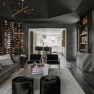 A modern living room with dark walls exuding sophisticated glamour, featuring a geometric chandelier, a wine rack, a gray couch, two black stools, and a coffee table. An open doorway reveals a bright kitchen in the background.