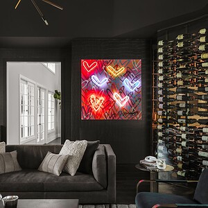 Modern living room with dark decor, exuding sophisticated glamour, featuring a neon heart art piece on the wall, a large wine rack, a grey sofa with cushions, two navy chairs, and a coffee table with a tray. Natural light streams in.
