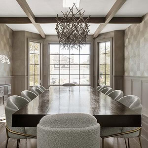 A modern dining room exudes sophisticated glamour, featuring a long, dark wooden table with eight upholstered chairs. A large geometric chandelier hangs above, and natural light streams in through the windows at the far end.