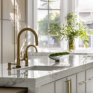 Modern kitchen with white marble countertops, brass fixtures, and wooden cabinetry exuding sophisticated glamour. A vase with green foliage is placed by a sunlit window. Black mesh pendant lights hang above the island.