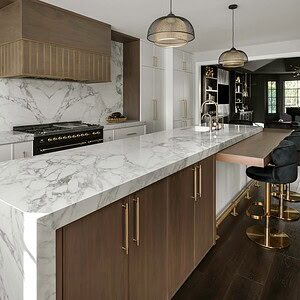 Modern kitchen featuring a marble island with wooden cabinetry, black and gold bar stools, pendant lights, and a sleek gas range with a wooden hood, exuding sophisticated glamour.