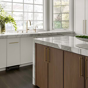 A modern kitchen exuding sophisticated glamour, featuring white cabinets with gold handles, a marble countertop, a large window above the sink, and wooden lower cabinets. Greenery is placed near the window.