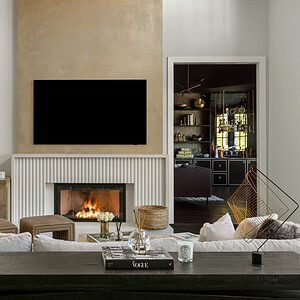 A modern living room exudes sophisticated glamour with a wall-mounted TV above a fireplace, surrounded by plush sofas and tasteful decor. An open door reveals a different, equally elegant room in the background.