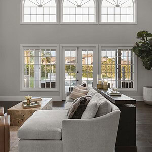 A modern living room showcasing sophisticated glamour with a large white sectional sofa, two brown ottomans, a wooden coffee table, a tall potted plant, and expansive windows and doors letting in natural light.