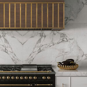 A stylish kitchen exuding sophisticated glamour features a marble backsplash, a dark gas range with gold detailing, a wooden range hood with vertical stripes, and a bowl containing two eggplants on the counter.