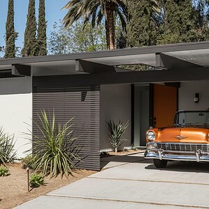 A classic Eichler Orange car is parked under the shade of a mid-century modern house carport surrounded by trees and desert plants.