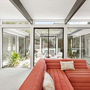 A modern living room with a red sectional sofa and large glass windows overlooking an indoor courtyard filled with plants. The space features a minimalist design with white walls, exposed beams, and a touch of Eichler Orange to add warmth and vibrance.