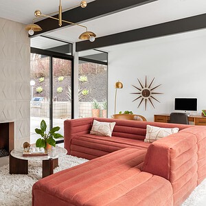 Modern living room with a large angular pink sofa, geometric patterned fireplace, glass coffee table, and contemporary chandelier. A desk with a computer is in the background, and large windows let in natural light. Accents of Eichler Orange add a vibrant touch to the overall decor.