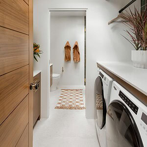 A laundry room with a skylight, washer, dryer, Eichler Orange wooden cabinets, and a doorway leading to a bathroom with two towels hanging on the wall and a geometric-patterned rug on the floor.