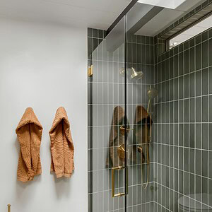 A modern bathroom with a glass-enclosed shower, green tiling, and brass fixtures. Two brown towels hang on the wall next to the shower, complemented by an Eichler Orange accent that adds a pop of color. A patterned bathmat lies on the floor.